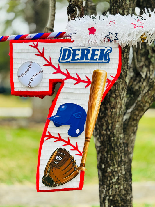 Handmade baseball piñata shaped like a number, decorated with red stitching, baseball glove, bat, and helmet details. Personalized with custom name ‘Derek’ and finished with bright red borders. Available in 26- or 36-inch sizes from Wild Corazon Designs.